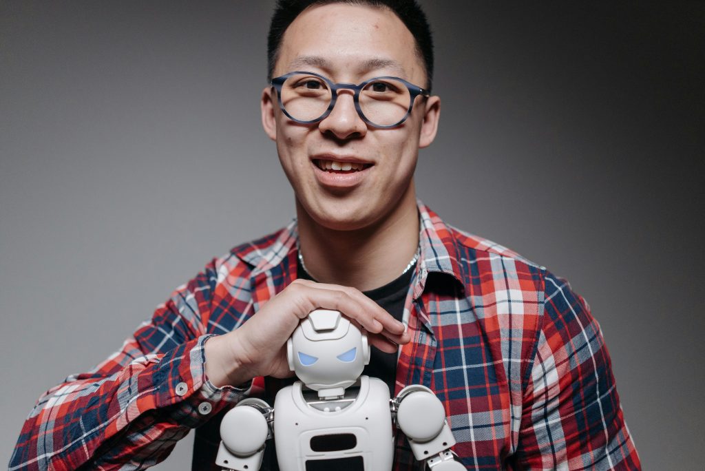 Asian man smiling, holding a small toy robot during a studio shoot. Indoors, with eyeglasses and plaid shirt.