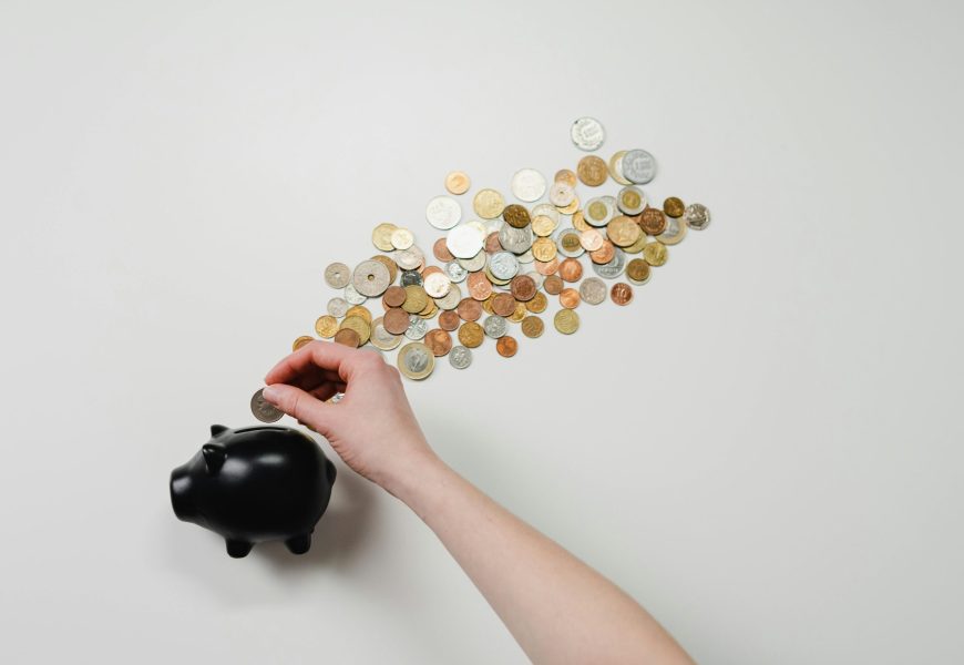 Person placing coin into a black piggy bank with scattered coins, symbolizing savings and finance.