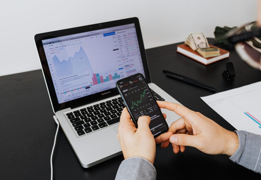 Person analyzing stock market data on a laptop and smartphone indoors.