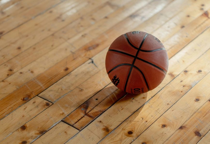 A single basketball resting on a polished wooden gym floor, casting a shadow.