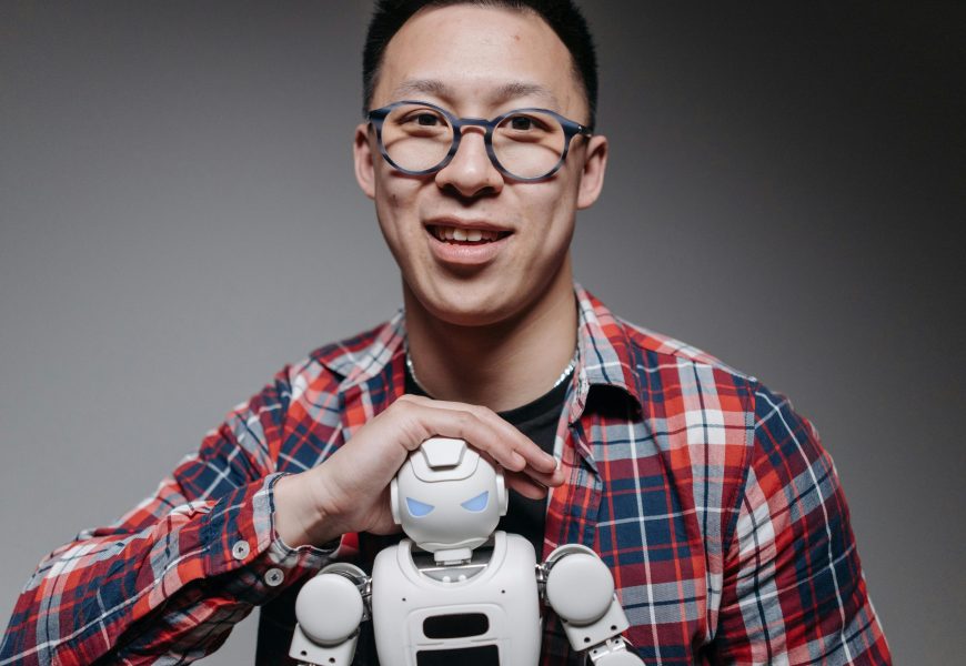 Asian man smiling, holding a small toy robot during a studio shoot. Indoors, with eyeglasses and plaid shirt.