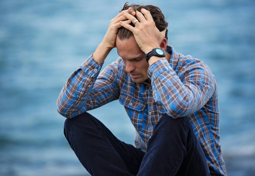 A man in a plaid shirt sits by the water looking distressed, symbolizing stress.